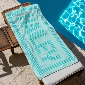 Puede incluir: Una toalla de playa azul claro con el nombre "HALEY" impreso en una fuente retro. La toalla tiene un borde y texto turquesa. Est&aacute; colocada en una tumbona de madera junto a una piscina.