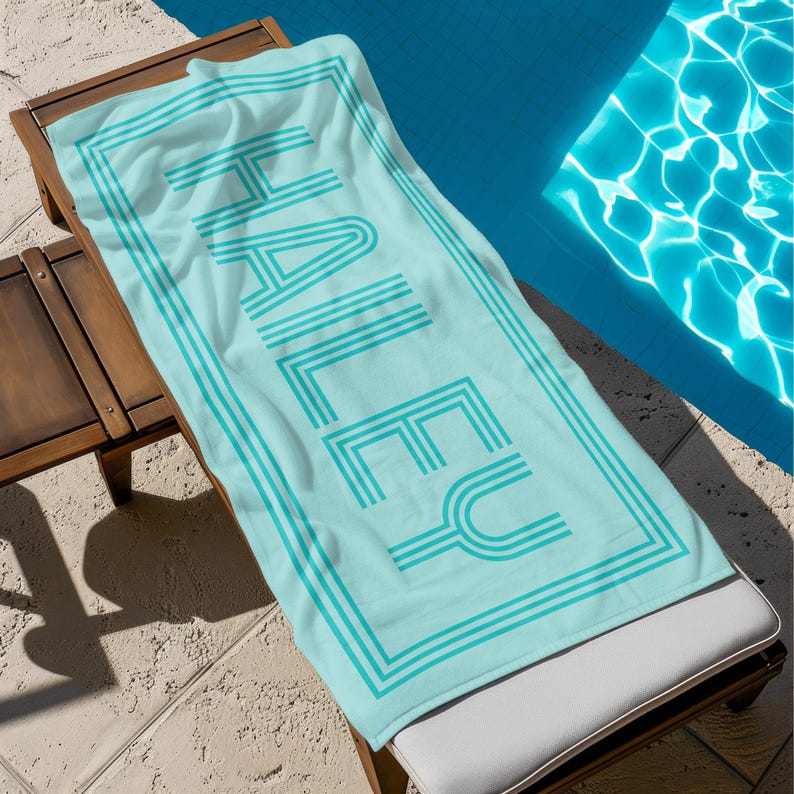 Puede incluir: Una toalla de playa azul claro con el nombre "HALEY" impreso en una fuente retro. La toalla tiene un borde y texto turquesa. Est&aacute; colocada en una tumbona de madera junto a una piscina.