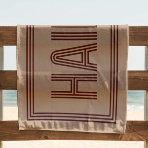 Puede incluir: Una toalla de playa beige con la palabra "HAI" en letras de bloque rojo oscuro. La toalla est&aacute; colgada en una valla de madera con una playa y el oc&eacute;ano de fondo. La toalla tiene un borde rojo oscuro.