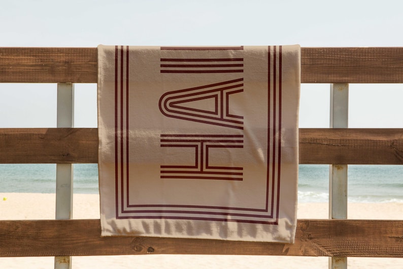 Puede incluir: Una toalla de playa beige con la palabra "HAI" en letras de bloque rojo oscuro. La toalla est&aacute; colgada en una valla de madera con una playa y el oc&eacute;ano de fondo. La toalla tiene un borde rojo oscuro.