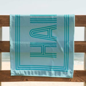 Puede incluir: Una toalla de playa azul claro con la palabra "HAI" en turquesa, con una fuente retro. La toalla est&aacute; colgada en una valla de madera con la playa y el mar al fondo. La toalla tiene rayas turquesas y un borde turquesa.