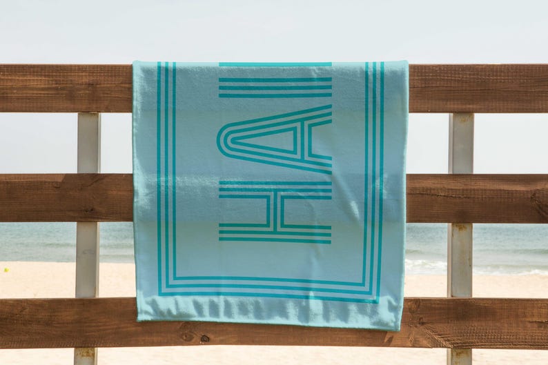 Puede incluir: Una toalla de playa azul claro con la palabra "HAI" en turquesa, con una fuente retro. La toalla est&aacute; colgada en una valla de madera con la playa y el mar al fondo. La toalla tiene rayas turquesas y un borde turquesa.