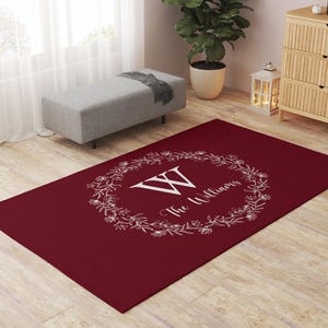 May include: A rectangular burgundy rug with a white floral wreath design. The wreath surrounds the letter "W" and the words "The Williams" in a script font. The rug is placed on a light wood floor.