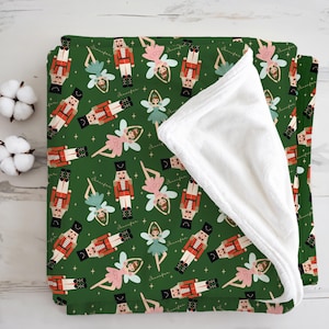 May include: A green blanket with a repeating pattern of nutcrackers and fairies. The nutcrackers are in red and black uniforms, and the fairies are in pink and teal dresses. The blanket has a white, fluffy underside.