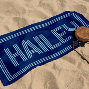 Puede incluir: Una toalla de playa azul marino con el nombre "HAILEY" en azul claro, delineado con un borde doble. Una bolsa redonda tejida y gafas de sol descansan sobre la toalla, todo sobre una playa arenosa.