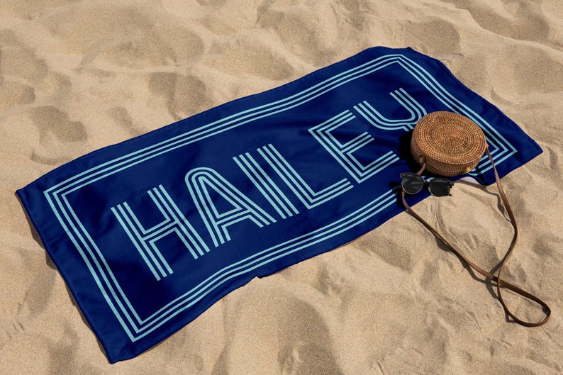 Puede incluir: Una toalla de playa azul marino con el nombre "HAILEY" en azul claro, delineado con un borde doble. Una bolsa redonda tejida y gafas de sol descansan sobre la toalla, todo sobre una playa arenosa.