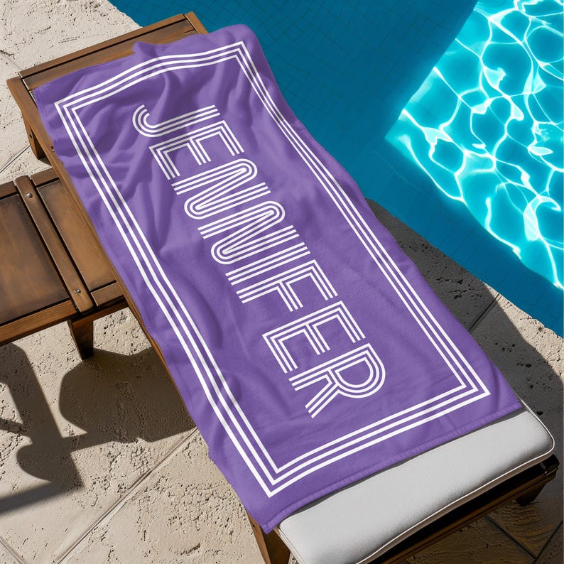 Puede incluir: Una toalla de playa morada con el nombre "JENNIFER" impreso en blanco, dentro de un borde blanco. La toalla est&aacute; colocada sobre una tumbona de madera junto a una piscina.