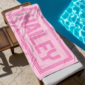 Puede incluir: Una toalla de playa rosa con el nombre "HALEY" impreso en una fuente retro. La toalla tiene un borde rosa y est&aacute; sobre una tumbona de madera junto a una piscina.