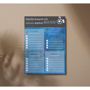 May include: A blue and white notepad with German text, including "Hierfür brauche ich meinen starken MANN!" and sections for lists. The notepad features illustrations of a superhero and muscular arms. It is mounted on a beige wall.