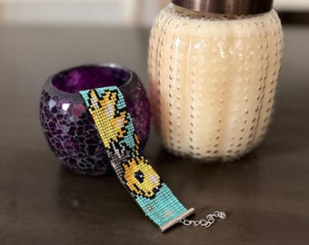 May include: A beaded bracelet featuring a floral pattern in yellow, black, and turquoise. The bracelet is made of small beads and has a silver clasp. A purple candle holder and a glass jar with a copper lid are in the background.