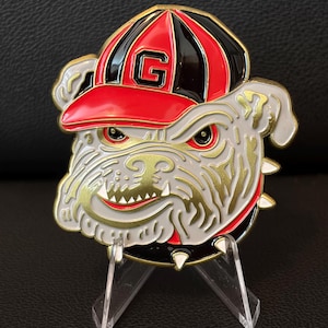 Puede incluir: Una moneda de metal con un bulldog que lleva una gorra de béisbol roja, negra y blanca con un contorno dorado y la letra "G". El bulldog tiene una expresión severa, detalles dorados y un collar con púas. Se muestra en un soporte transparente.