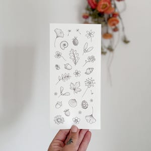 May include: A rectangular card with detailed black line drawings of natural elements such as leaves, flowers, and berries. The illustrations are arranged on a white background. The card is held by a hand, with a tattoo visible on the thumb.