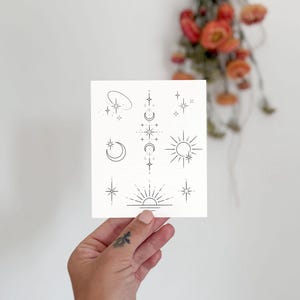 以下が含まれることがあります： 星、月、太陽の光線など、ミニマリストの天体デザインが施された白いカード。イラストは黒インクです。カードは小さなタトゥーのある手で持たれています。背景には乾燥したオレンジ色の花があります。