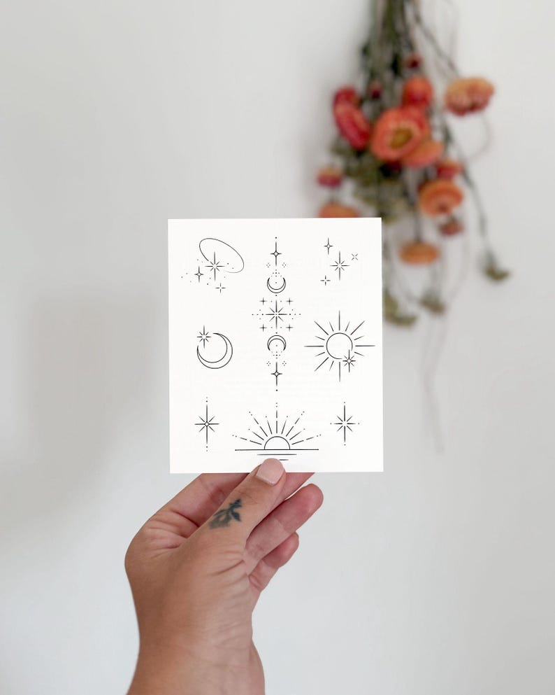 May include: A white card featuring minimalist celestial designs, including stars, moons, and a sun with rays. The illustrations are in black ink. The card is held in a hand with a small tattoo. Dried orange flowers are in the background.