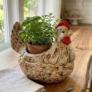 May include: A ceramic chicken-shaped planter with a brown and cream mottled glaze, featuring a red comb and wattle. The planter holds a small terracotta pot with green herbs. A folded beige linen napkin and a small silver spoon sit nearby.