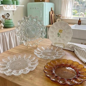 May include: A collection of four round serving platters designed for eggs, including three clear glass plates with textured patterns and one white ceramic plate featuring a yellow rose illustration in the centre, all arranged on a wooden surface.