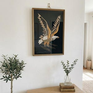 May include: A rectangular framed wall art piece featuring a detailed illustration of an eagle in flight. The eagle is rendered in shades of gold, brown, and white against a solid black background, enclosed within a simple, thin wooden frame.