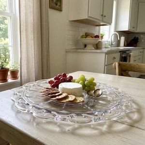May include: A clear glass serving platter with a decorative scalloped edge, holding a round wheel of white cheese, a cluster of red grapes, a cluster of green grapes, a stack of round crackers, and two small silver-toned metal spoons.