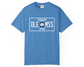 May include: A sky blue t-shirt with a white license plate design. The license plate reads "MISSISSIPPI" at the top, "OLE MSS" in the centre with a flower graphic, and "OXFORD" at the bottom. The shirt is a short-sleeved crew neck.