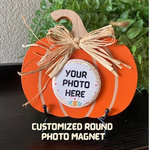 May include: An orange wooden pumpkin decoration with a raffia bow and a round magnet that says "YOUR PHOTO HERE." The text "CUSTOMIZED ROUND PHOTO MAGNET" is below the pumpkin.