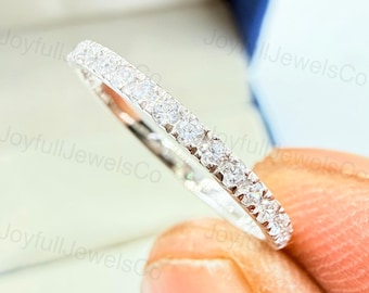 Puede incluir: Un anillo plateado con una fila de pequeñas piedras preciosas transparentes engastadas a lo largo de la banda. El anillo se sostiene entre dos dedos, con el foco en las piedras brillantes. Es una joya.