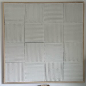 Może przedstawiać: Duże, kwadratowe, teksturowane dzieło sztuki ściennej z jasną drewnianą ramą. Grafika podzielona jest na szesnaście kwadrat&oacute;w, każdy z unikalnym pionowym lub poziomym żebrowanym wzorem. Kolor jest neutralny, białawy.