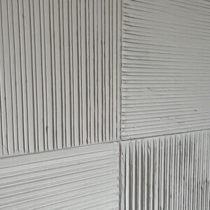 Może przedstawiać: Zbliżenie teksturowanego, kremowobiałego panelu ściennego. Panel podzielony jest na cztery sekcje, każda z unikalnym wzorem pionowych i poziomych grzbiet&oacute;w. Grzbiety tworzą grę światła i cienia.