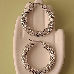 May include: Silver hoop earrings with a beaded design, displayed on a white ceramic hand. The earrings are circular and have a textured surface. The background is a pale green colour, providing a contrast to the earrings.