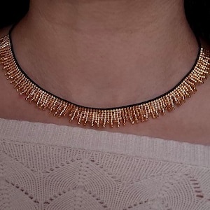 Pu&ograve; includere: Una collana di perline dorate con un cordino nero. La collana presenta un design a frange con piccole perline dorate. La collana &egrave; indossata sopra un maglione bianco strutturato.