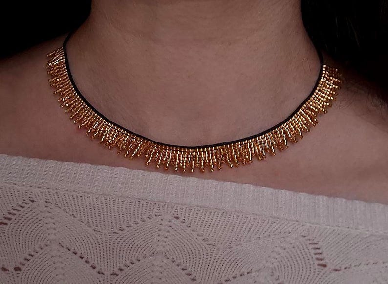 Pu&ograve; includere: Una collana di perline dorate con un cordino nero. La collana presenta un design a frange con piccole perline dorate. La collana &egrave; indossata sopra un maglione bianco strutturato.