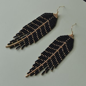 May include: A pair of black beaded feather earrings with gold accents. The earrings have a gold hook and are made of small black beads with gold beads forming a central vein and side veins. The earrings are on a light green surface.