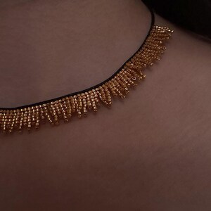 Pu&ograve; includere: Una collana di perline dorate con un cordoncino nero. La collana presenta una frangia di piccole perline dorate che pendono dal cordoncino. Le perline sono di un caldo colore dorato e la collana &egrave; mostrata su uno sfondo color carne.