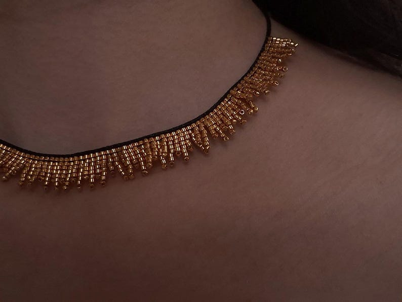Pu&ograve; includere: Una collana di perline dorate con un cordoncino nero. La collana presenta una frangia di piccole perline dorate che pendono dal cordoncino. Le perline sono di un caldo colore dorato e la collana &egrave; mostrata su uno sfondo color carne.