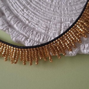 Pu&ograve; includere: Una collana di perline dorate con una fascia nera. La collana presenta file di piccole perline dorate che pendono, creando un effetto frangia. La collana &egrave; esposta su una superficie bianca strutturata su uno sfondo verde chiaro.