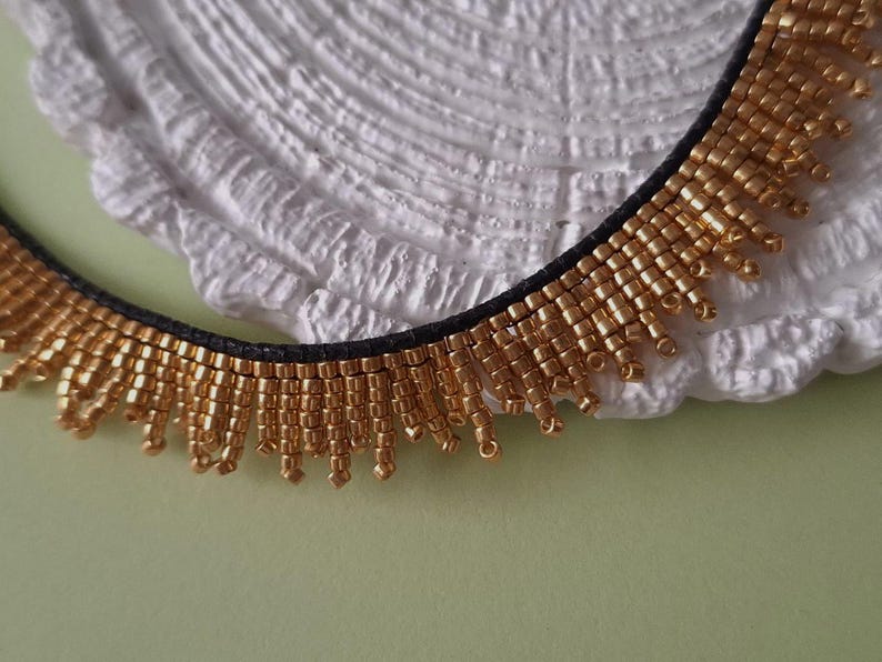Pu&ograve; includere: Una collana di perline dorate con una fascia nera. La collana presenta file di piccole perline dorate che pendono, creando un effetto frangia. La collana &egrave; esposta su una superficie bianca strutturata su uno sfondo verde chiaro.