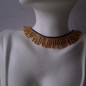 Pu&ograve; includere: Una collana di perline dorate con base nera &egrave; esposta su un manichino bianco. La collana presenta un design a frange con piccole perline dorate che pendono dalla base. Le perline sono di un caldo colore oro.