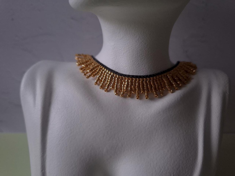 Pu&ograve; includere: Una collana di perline dorate con base nera &egrave; esposta su un manichino bianco. La collana presenta un design a frange con piccole perline dorate che pendono dalla base. Le perline sono di un caldo colore oro.