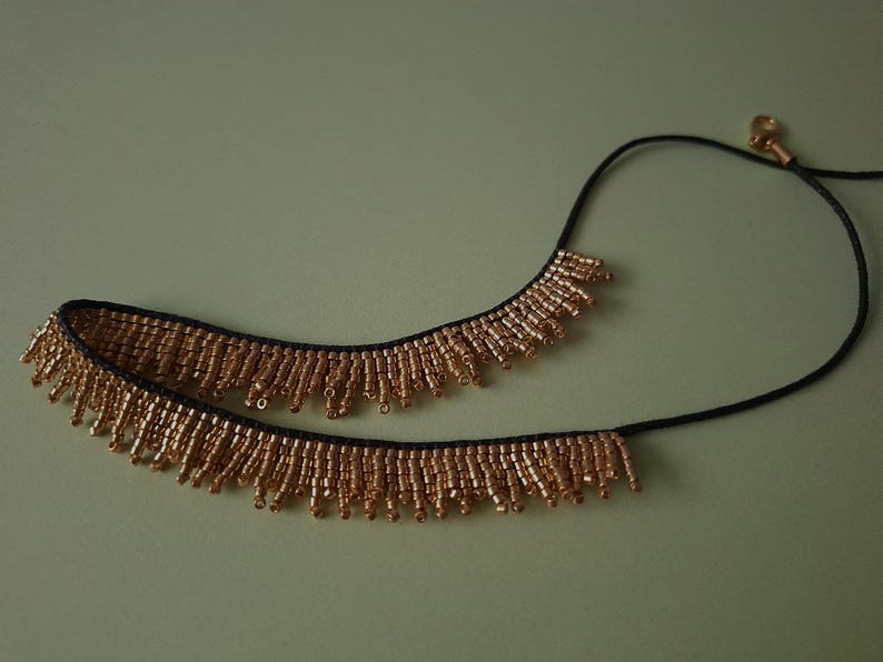 Pu&ograve; includere: Una collana di perline dorate con un cordino nero. La collana presenta una frangia di piccole perline dorate. La collana &egrave; su uno sfondo verde chiaro. &Egrave; un gioiello.