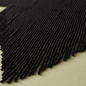 May include: Close-up of a black beaded fringe. The fringe is made of small, round beads strung together, creating a textured surface. The fringe hangs from a woven base, with the individual strands of beads ending in a slightly thicker, elongated shape.