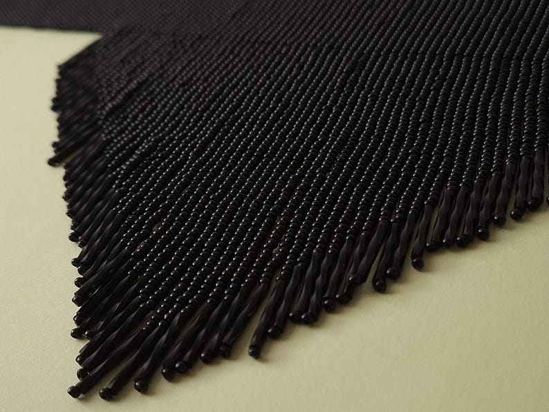 May include: Close-up of a black beaded fringe. The fringe is made of small, round beads strung together, creating a textured surface. The fringe hangs from a woven base, with the individual strands of beads ending in a slightly thicker, elongated shape.