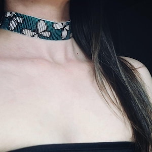 Könnte beinhalten: Ein tealfarbenes Perlen-Choker-Halsband mit einem Blumenmuster in Weiß und Schwarz. Das Choker-Halsband wird um den Hals getragen. Nahaufnahme eines modischen Accessoires.