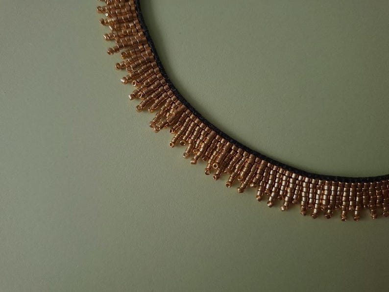 Pu&ograve; includere: Primo piano di una collana di perline dorate con retro nero. La collana presenta un design a frange con piccole perline dorate che pendono dal basso. Lo sfondo &egrave; di colore verde chiaro.