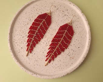 May include: A pair of red beaded feather earrings with gold-colored accents. The earrings are displayed on a speckled, off-white circular tray against a light green background. The earrings have a long, dangling design.
