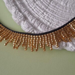 Pu&ograve; includere: Primo piano di una collana di perline dorate con retro nero. La collana ha una frangia di perline dorate. Lo sfondo &egrave; una superficie bianca strutturata e una superficie verde chiaro.