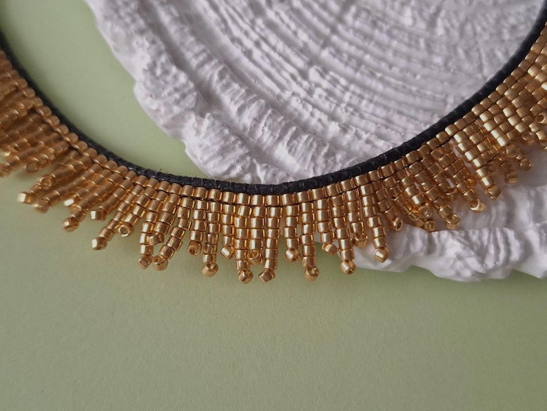 Pu&ograve; includere: Primo piano di una collana di perline dorate con retro nero. La collana ha una frangia di perline dorate. Lo sfondo &egrave; una superficie bianca strutturata e una superficie verde chiaro.