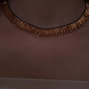 Pu&ograve; includere: Una collana di perline dorate con una fascia nera. La collana presenta un design a frange con piccole perline dorate appese alla fascia. La collana &egrave; indossata intorno al collo.