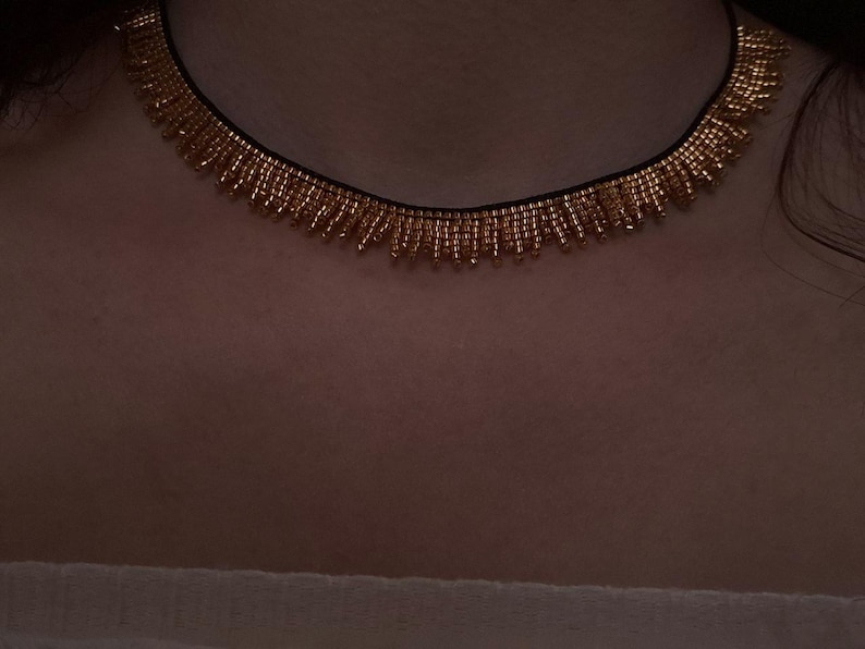 Pu&ograve; includere: Una collana di perline dorate con una fascia nera. La collana presenta un design a frange con piccole perline dorate appese alla fascia. La collana &egrave; indossata intorno al collo.