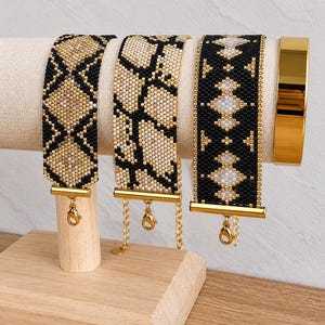 Pode incluir: Três pulseiras largas de missangas com padrões geométricos e de inspiração animal em tons de preto, dourado e creme, cada uma com terminais de metal dourado e fechos de lagosta, ao lado de uma pulseira rígida dourada lisa e polida.