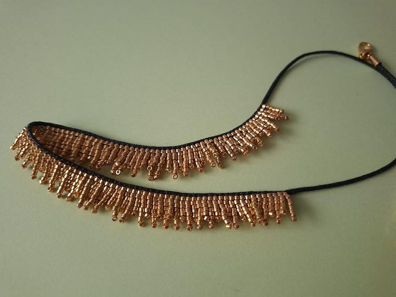 Pu&ograve; includere: Una collana di perline dorate con un cordino nero. La collana presenta una frangia di piccole perline dorate lungo il bordo inferiore. La collana &egrave; esposta su uno sfondo verde chiaro.