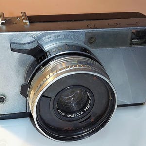 May include: Silver and black vintage Zorki 10 camera with a prominent lens. The camera has a rectangular body with a silver finish and a black lens. The lens has a silver and black ring. The camera has the text "Zorki 10" on the top.